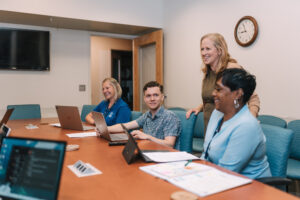 Christine Vanlandingham stands behind staff sitting at table during a meeting