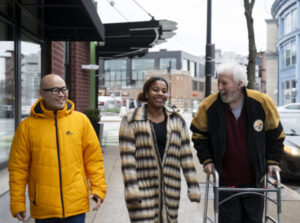 three people walk down a street together. one of them uses a walker
