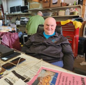 bald man in his 60s sits behind a counter at a thrift store