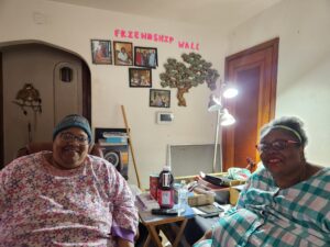 Two women sit in their living room smiling with "Friendship Wall" across the wall behind them.