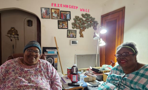 Two women sit in their living room smiling with "Friendship Wall" across the wall behind them.