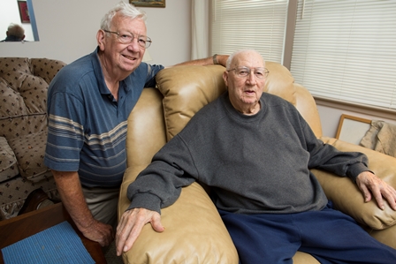 two men smiling at camera, one sitting in a chair and one kneeling behind