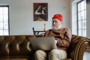 Man in red hat with beard sits on couch looking at laptop