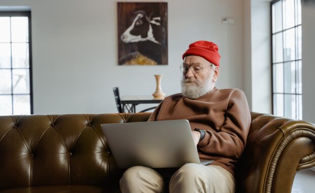 Man in red hat with beard sits on couch looking at laptop