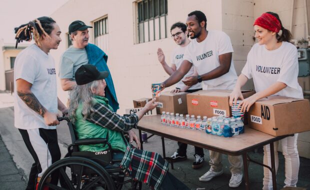 Scene depicts volunteers giving food items to a gentleman in a wheelchair